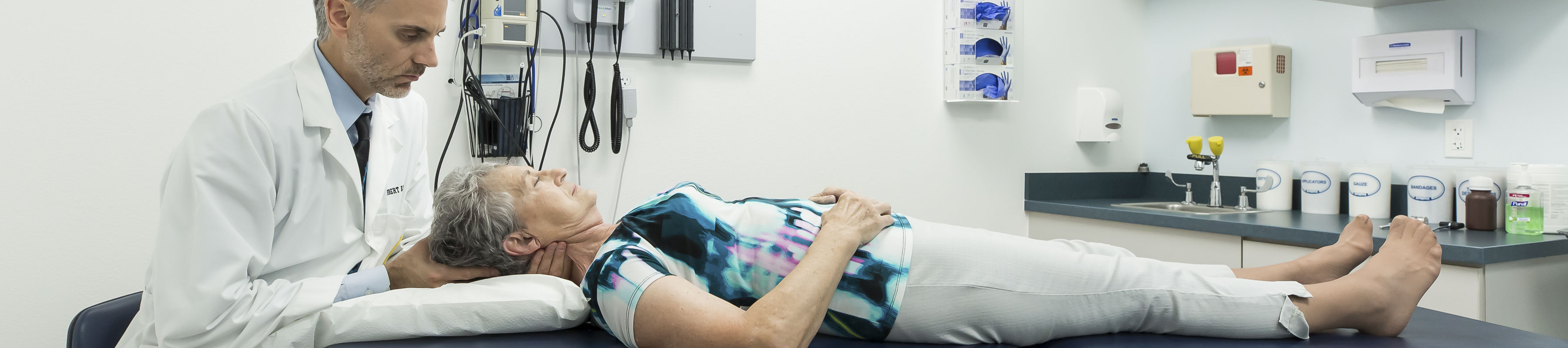 A patient lays on an exam table in a doctor office while a physician examines her neck