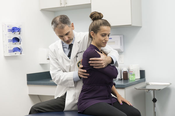 A patients sits on an exam table while a physician adjusts their shoulder